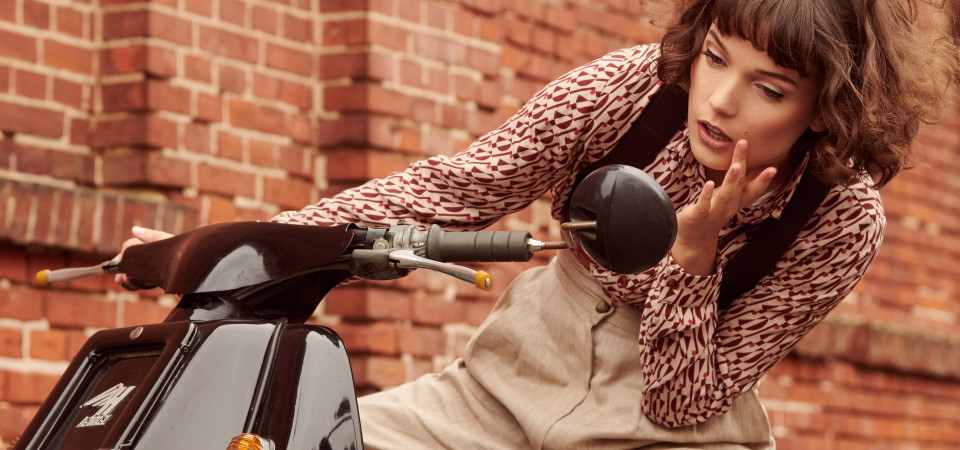 Eine Frau in einer Vintage-Bluse und einer hellen Marlenehose sitzt auf einem Motorroller