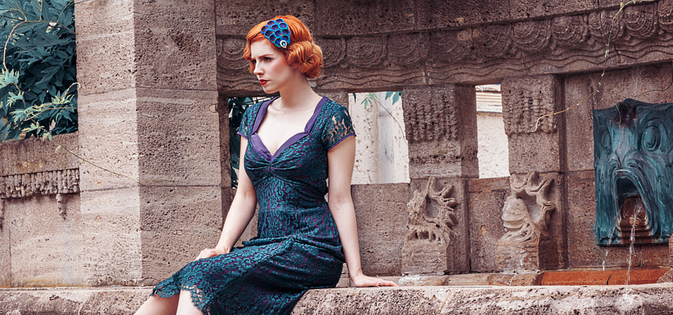 Eine Frau in einem grünen Spitzenkleid im Stil der 1930er Jahre sitzt an einem Brunnen