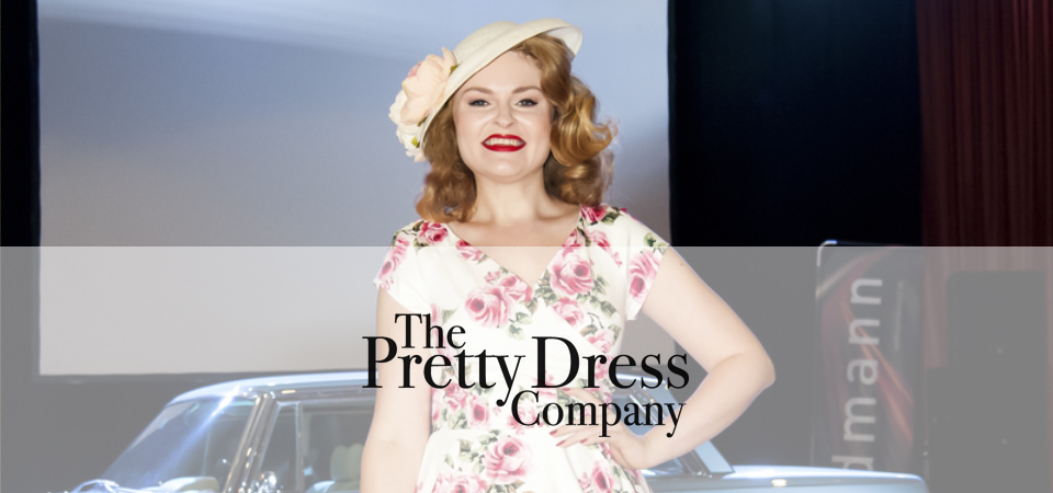 Eine Frau trägt ein geblümtes Kleid und einen Hut im 50er Jahre Stil, Logo der Marke Pretty Dress Company